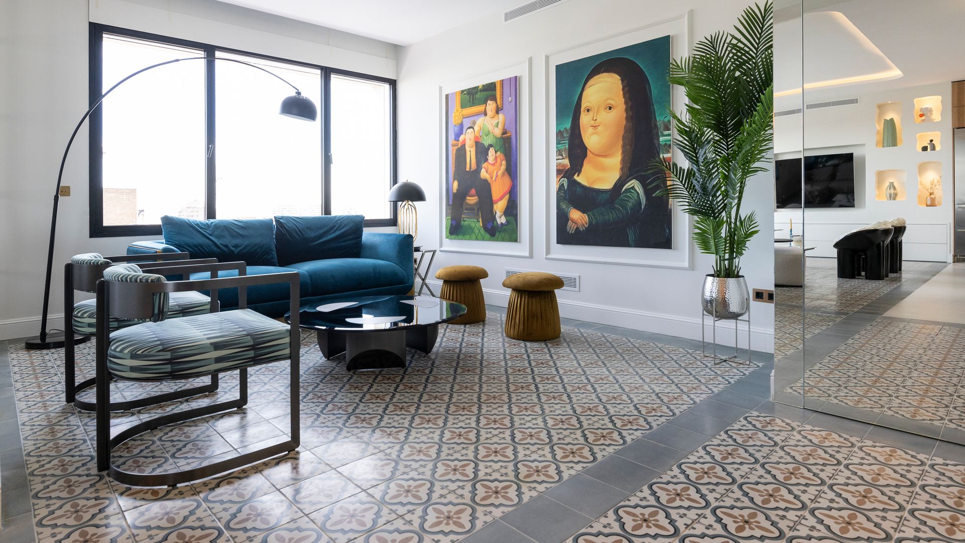 Salón con sofá azul, cuadros en la pared, suelo hidráulico, taburetes marrones, sillas, mesa de centro negra, lámpara de pie tipo arco
