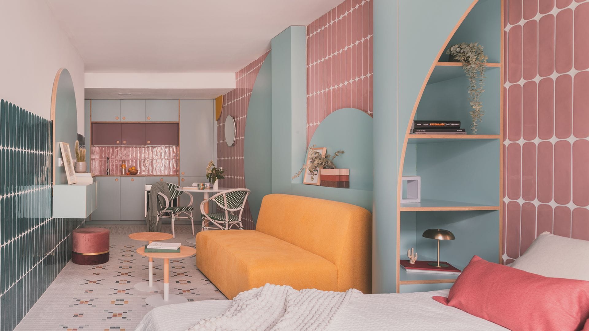 Espacio único con dormitorio, salón con sofá amarillo y cocina, paredes de azulejos rosas, y azules, bloques de madera azul de formas redondeadas