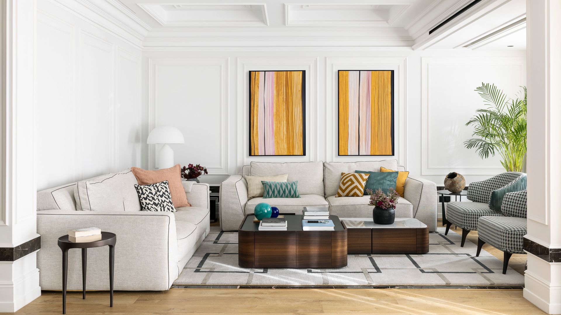 Salón con molduras decorativas, alfombra, cuadros en la pared. techo artesonado de color blanco, sofás blancos, mesas de centro de madera, butacas tapizadas con cuadros blancos y negros, cojines en el sofá de colores