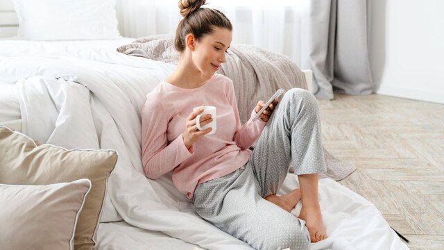 mujer joven mirando el móvil junto a la cama, tomando un café y con libros abiertos a sus pies