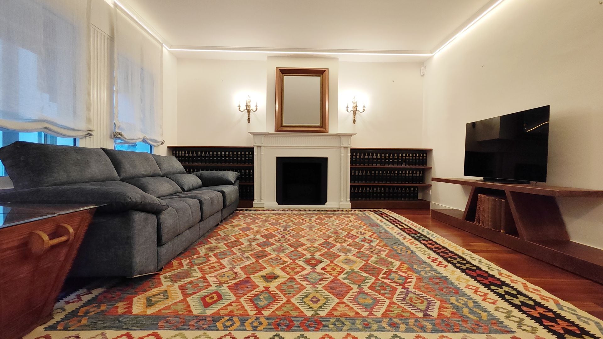 Salón con chimenea y alfombra de motivos geométricos, estores, mueble de televisión, apliques de pared, sofá de cuero negro, espejo sobre la chimenea