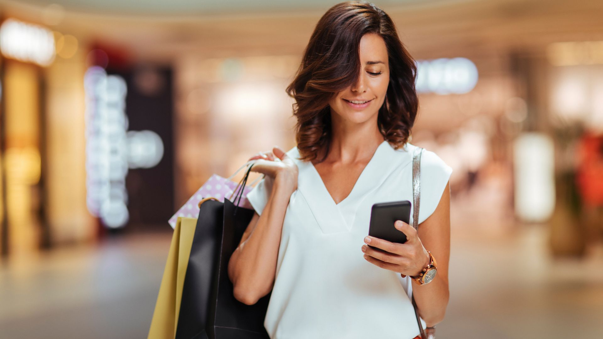 Mujer con teléfono móvil en el pasillo de un centro comercial, con ropa de entretiempo y bolsas en la otra mano.