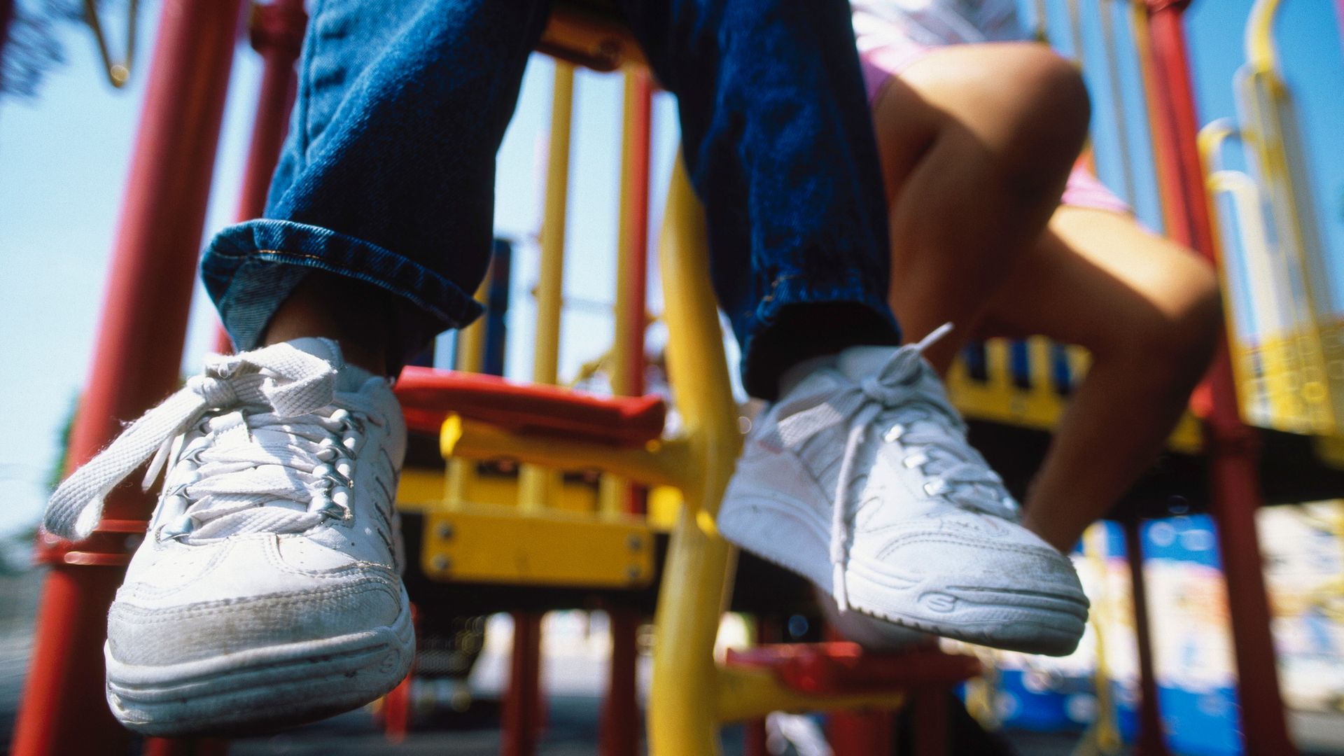 Niño jugando en un parque infantil al aire libre, está sentado sobre un columpio y solo se le ven los pantalones vaqueros y las zapatillas deportivas blancas.