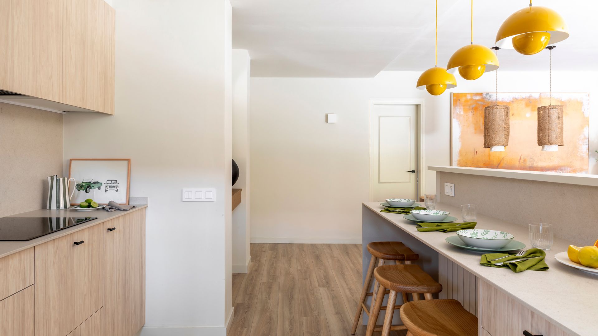 Cocina con isla con barra de desayunos, taburetes de madera, lámparas de techo de color amarillo, muebles de madera clara, suelo de madera oscuro, platos sobre la barra