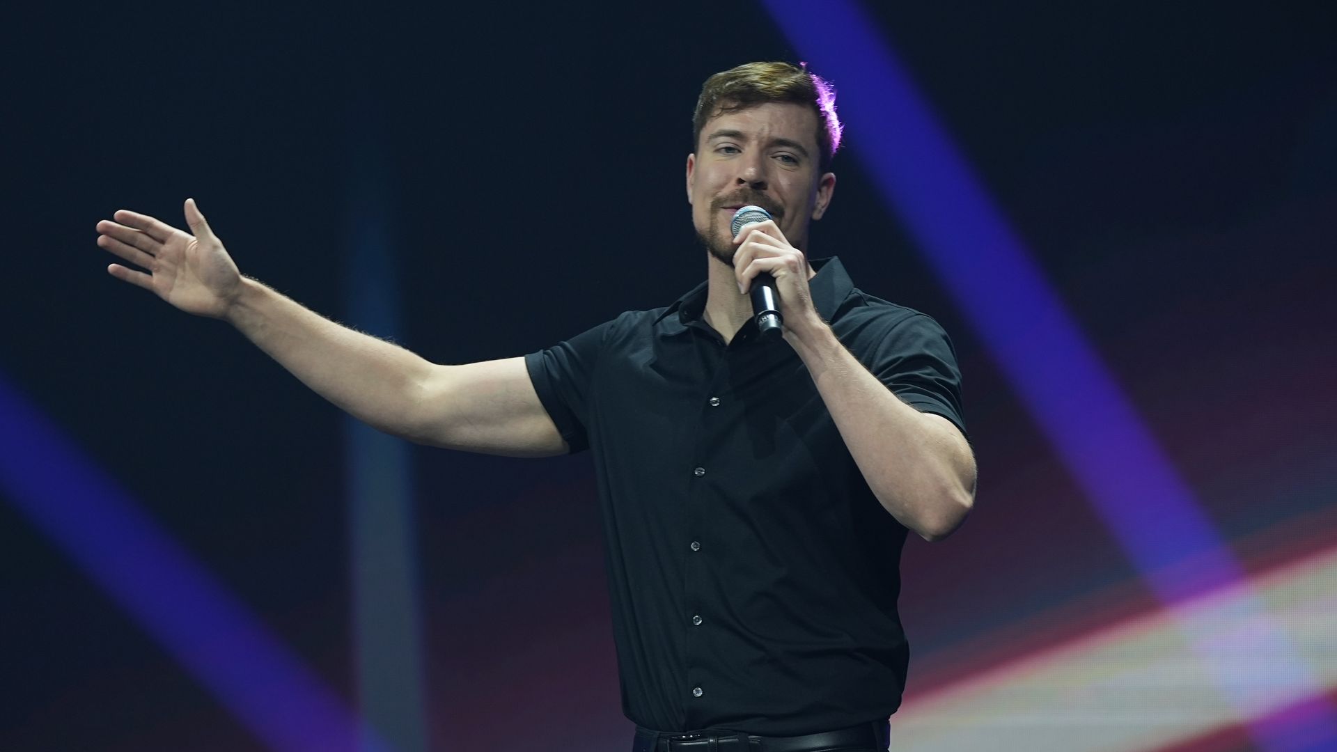MrBeast en un escenario, con micrófono, mano extendida, vestido de negro.