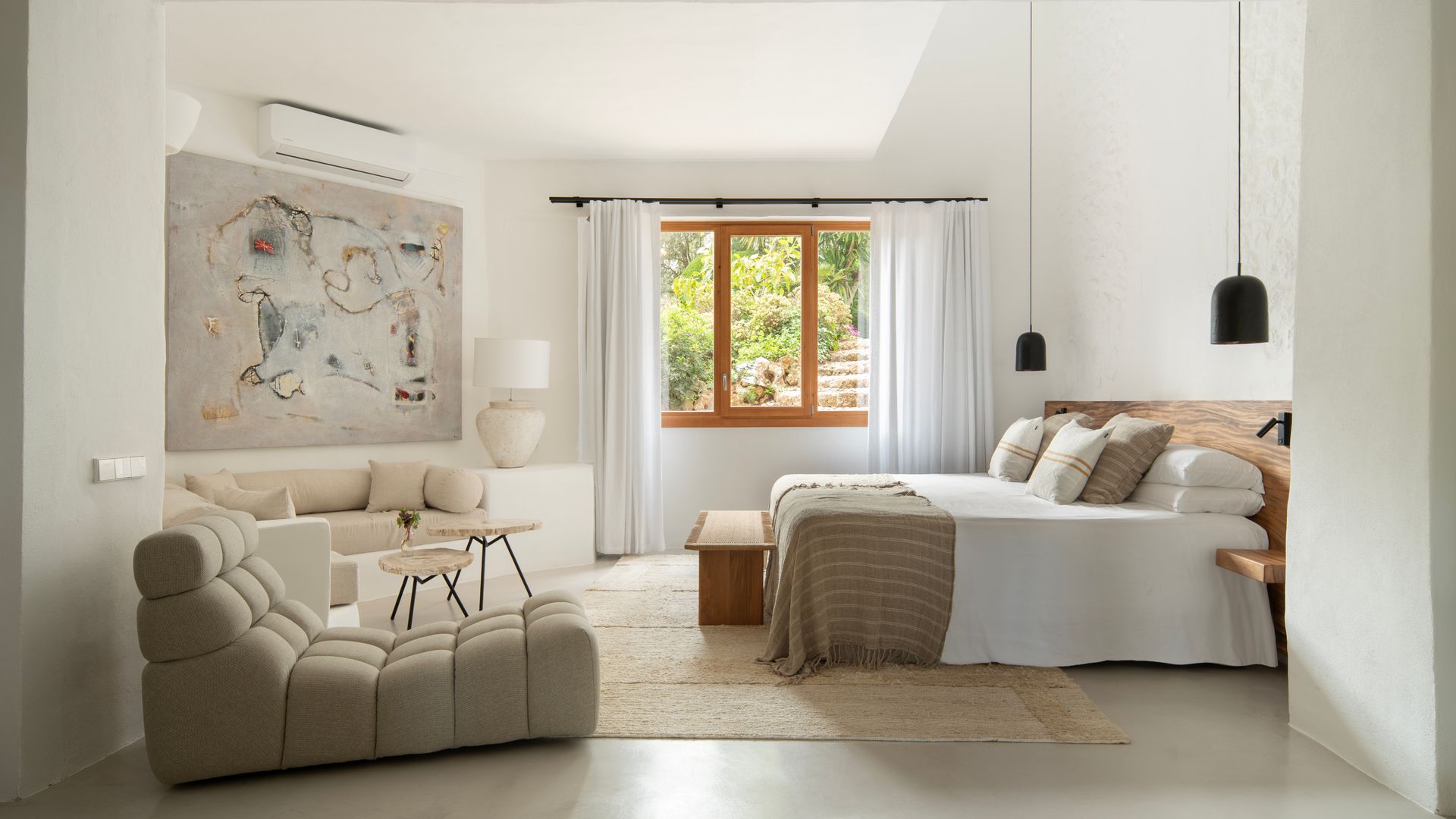 Dormitorio con banco de obra blanco, cabecero de madera, banqueta de madera a los pies, lámparas de techo negro a ambos lados de la cama, butaca moderna beige, ventanas con el marco de madera, cortinas blancas, alfombra