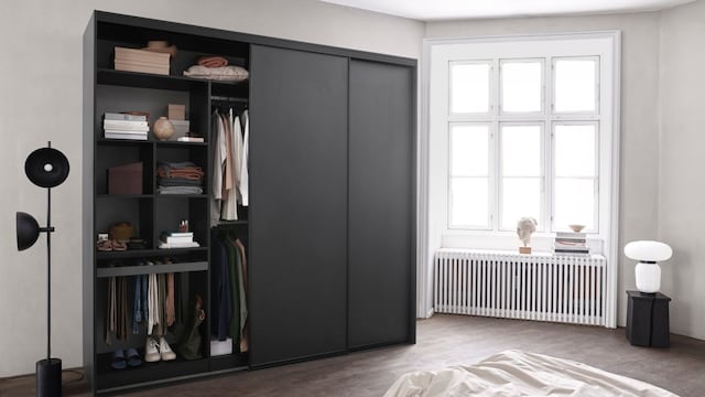 Dormitorio con armario con puertas negras, lámpara de pie, ventanal con radiador antiguo