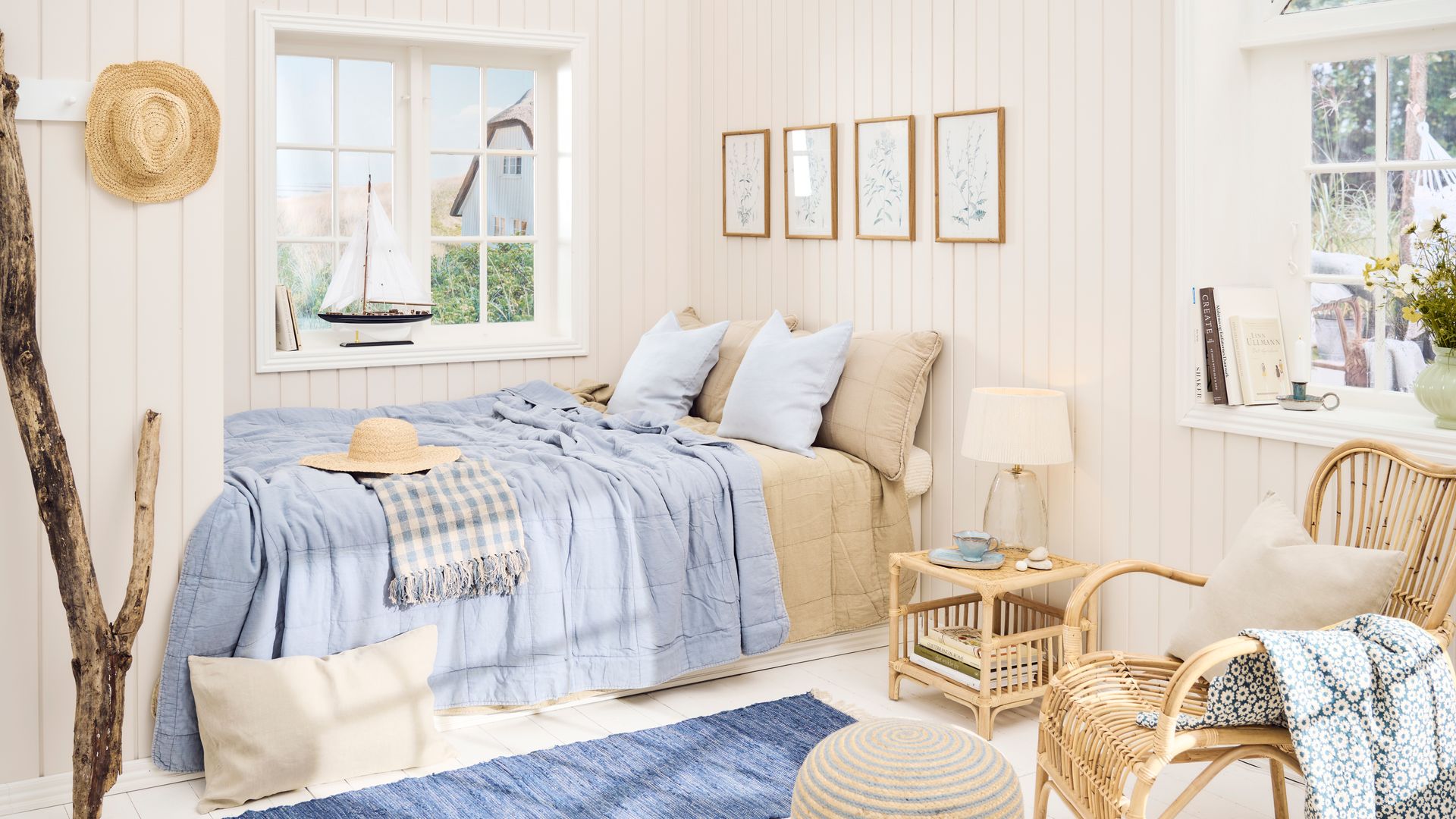 Dormitorio playero con lamas de madera, ropa de cama azul, cuadros en la pared, mesita de noche y butaca de fibras vegetales, alfombra, sombrero de paja colgado en perchero de pared, adorno de barco 