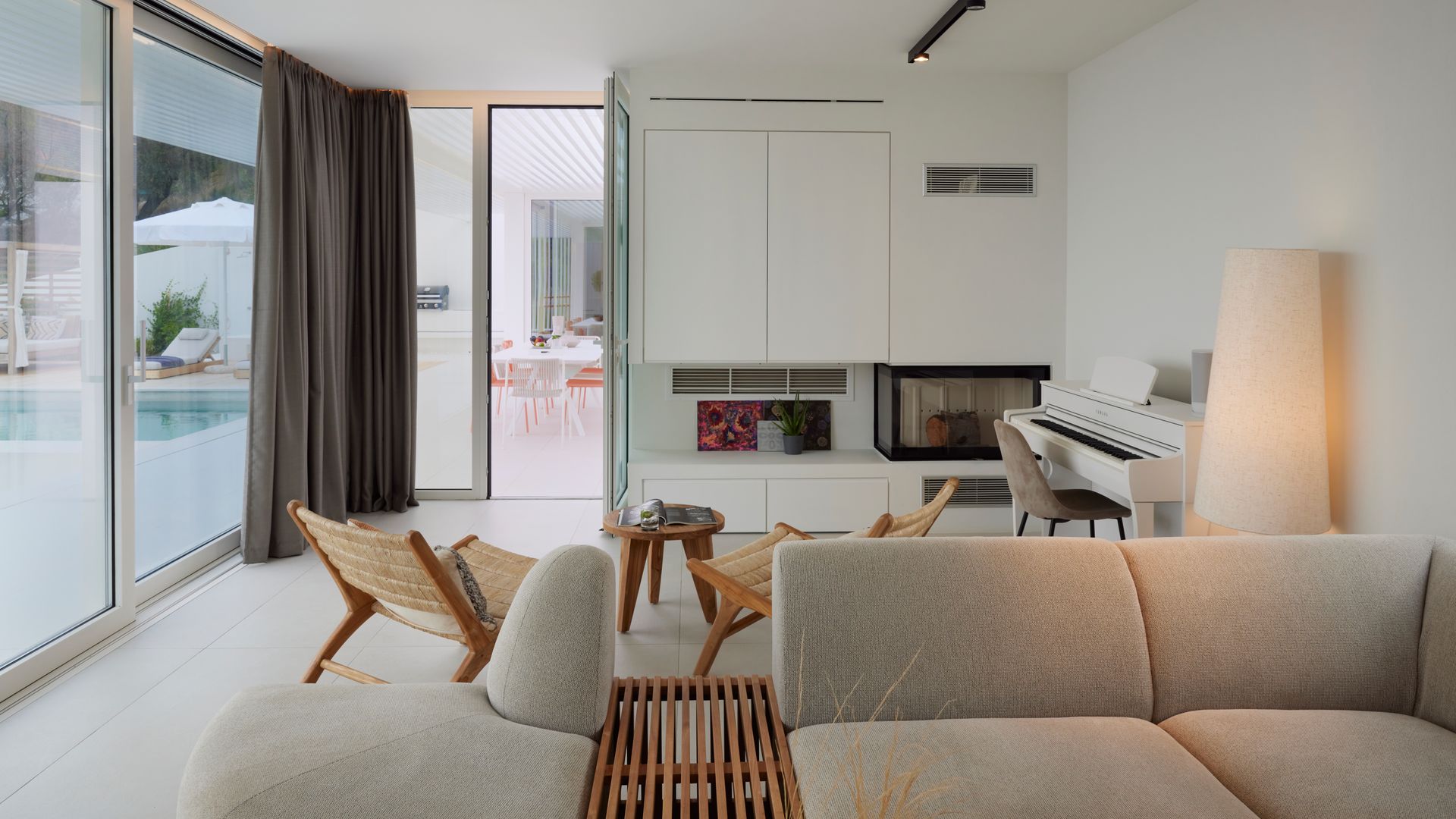 Salón con chimenea y piano blanco, salida a la piscina, sofá gris, rincón de lectura con dos butacas de fibras vegetales y mesita auxiliar 