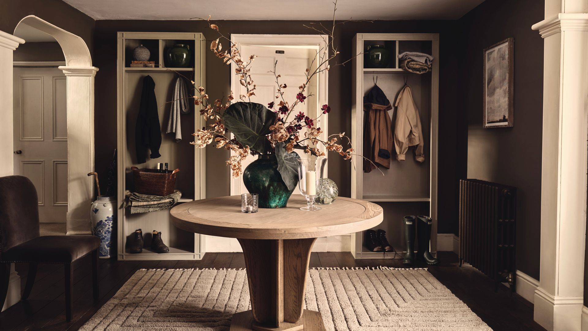 Recibidor con mesa redonda en el centro, jarrón con arreglo floral, alfombra beige, dos armarios abiertos, con zapatero, barra para colgar y baldas, 