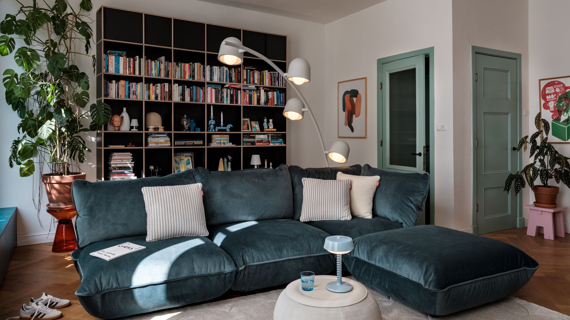 Salón con sofá e terciopelo en color azul marino, alfombra beige, mesa de centro redonda tapizada, lámpara de pie tras el sofá, librería con libros y adornos, puertas de paso de color verde claro, taburete rosa con maceta