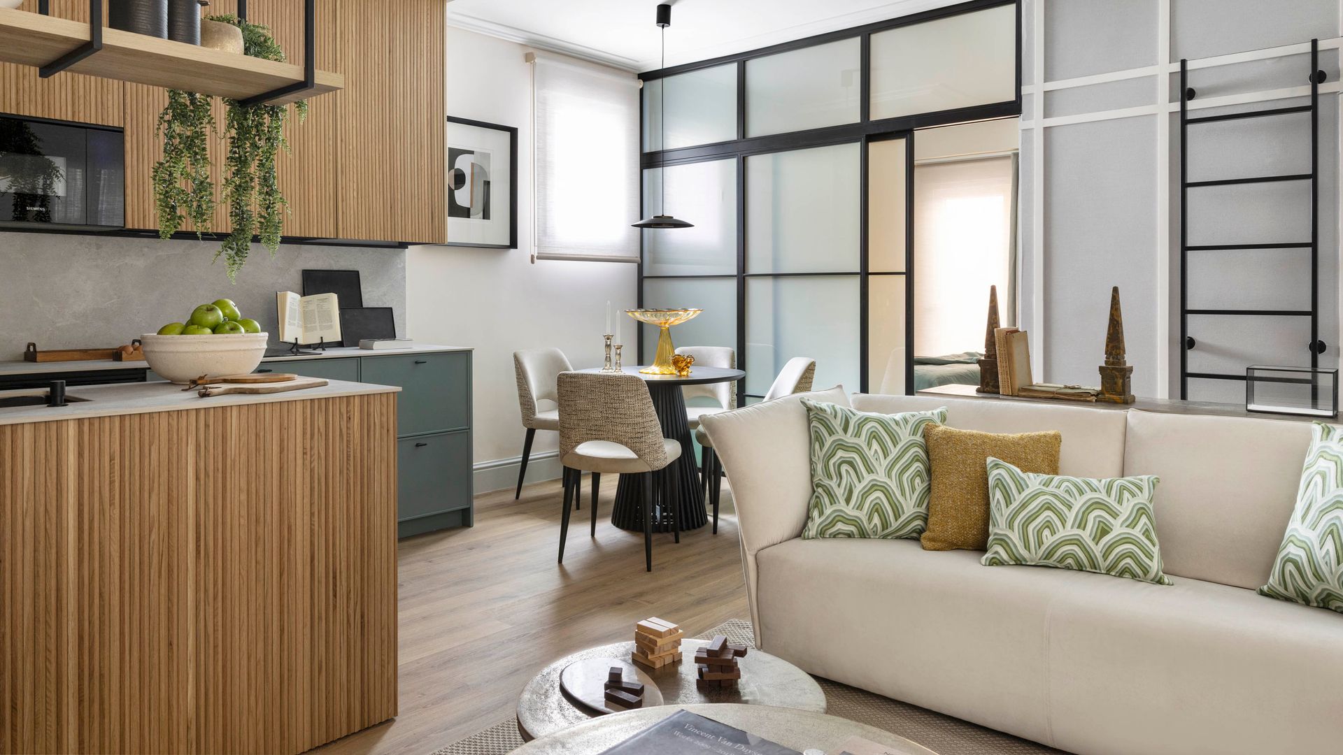 Salón comedor y cocina, paneles de cristal separando el dormitorio, sofá beige, isla, altillo almacenaje, isla de cocina con balda superior con cazuelas y plantas