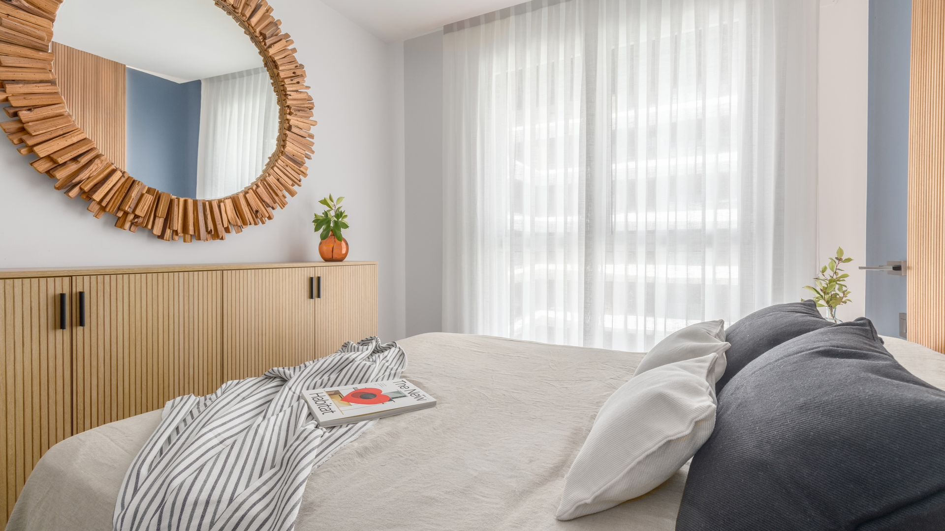 Dormitorio con espejo redondo, aparador de lamas de madera, cortinas blancas