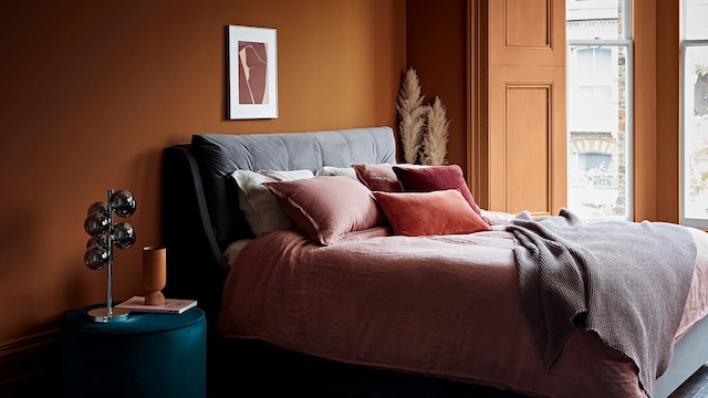 Dormitorio con paredes en color tierra, cabecero entelado, mesita de noche tipo puff y ropa de cama rosa