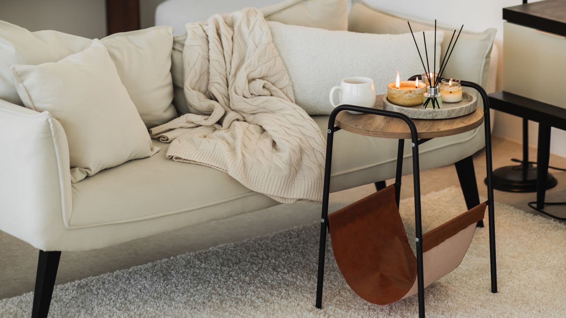 Salón con decoración minimalista y moderna, en tonos blancos y negros. Sofá de dos plazas con mesa auxilirar repleta de velas, mikados y una taza blanca. Al fondo, una estantería de madera pegada a la pared y también una lámpara de pie.