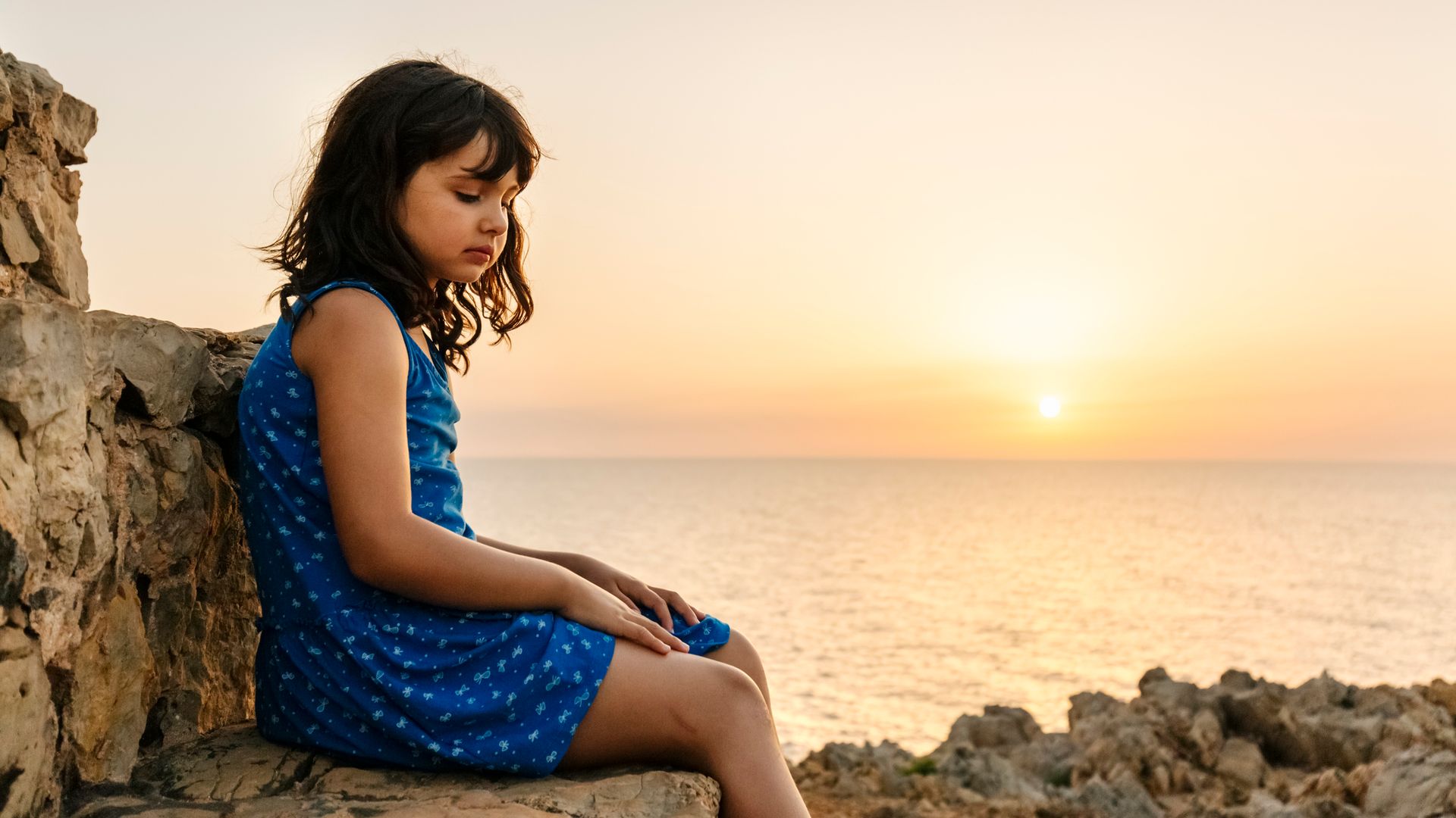 niña pensativa sentada junto al mar, con el sol en el horizonte