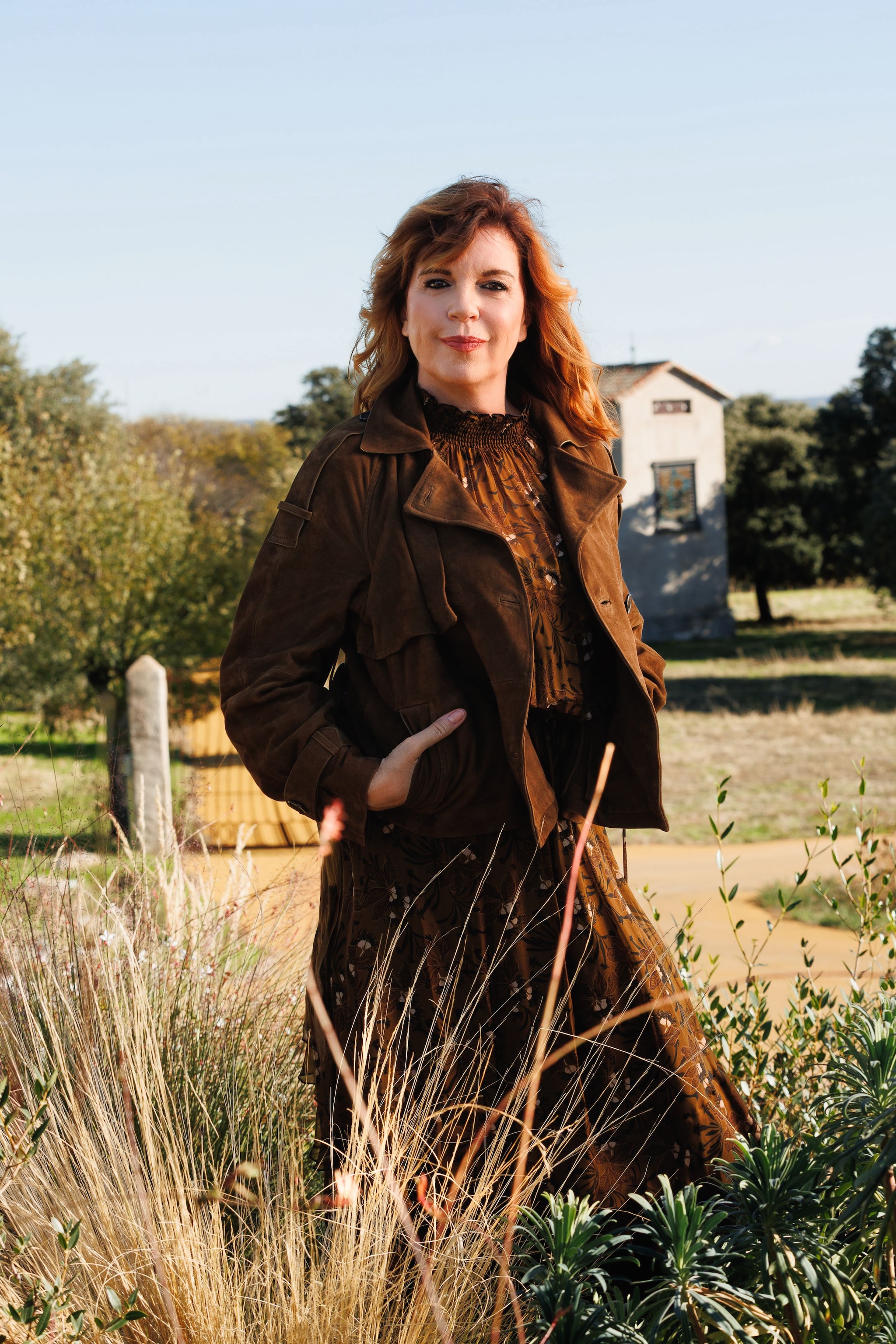 Mujer con cabello rizado, en un campo, con chaqueta y vestido marrones.