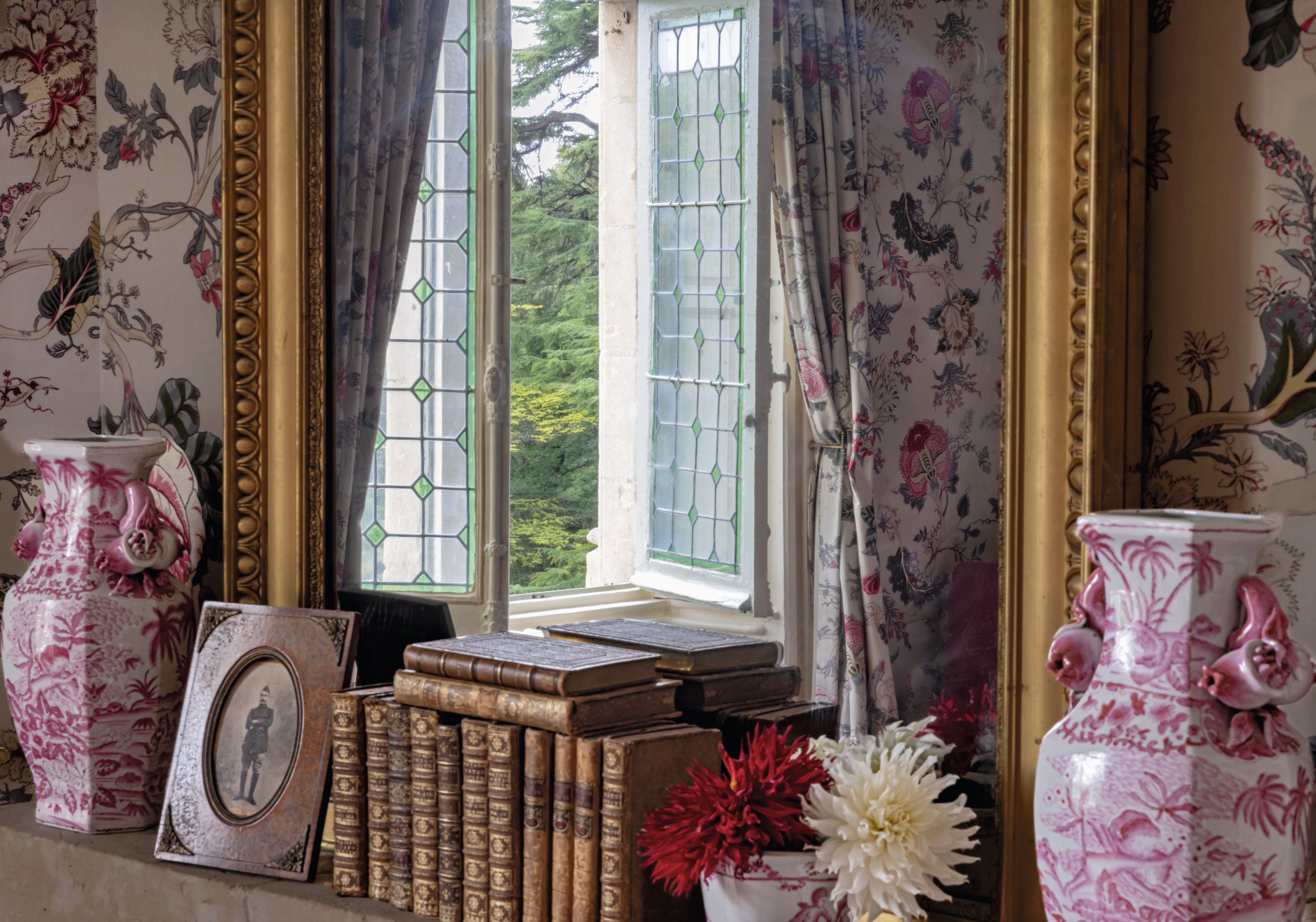 Espejo dorado con marco floral, mostrando una ventana con cortinas y un arreglo de libros y flores