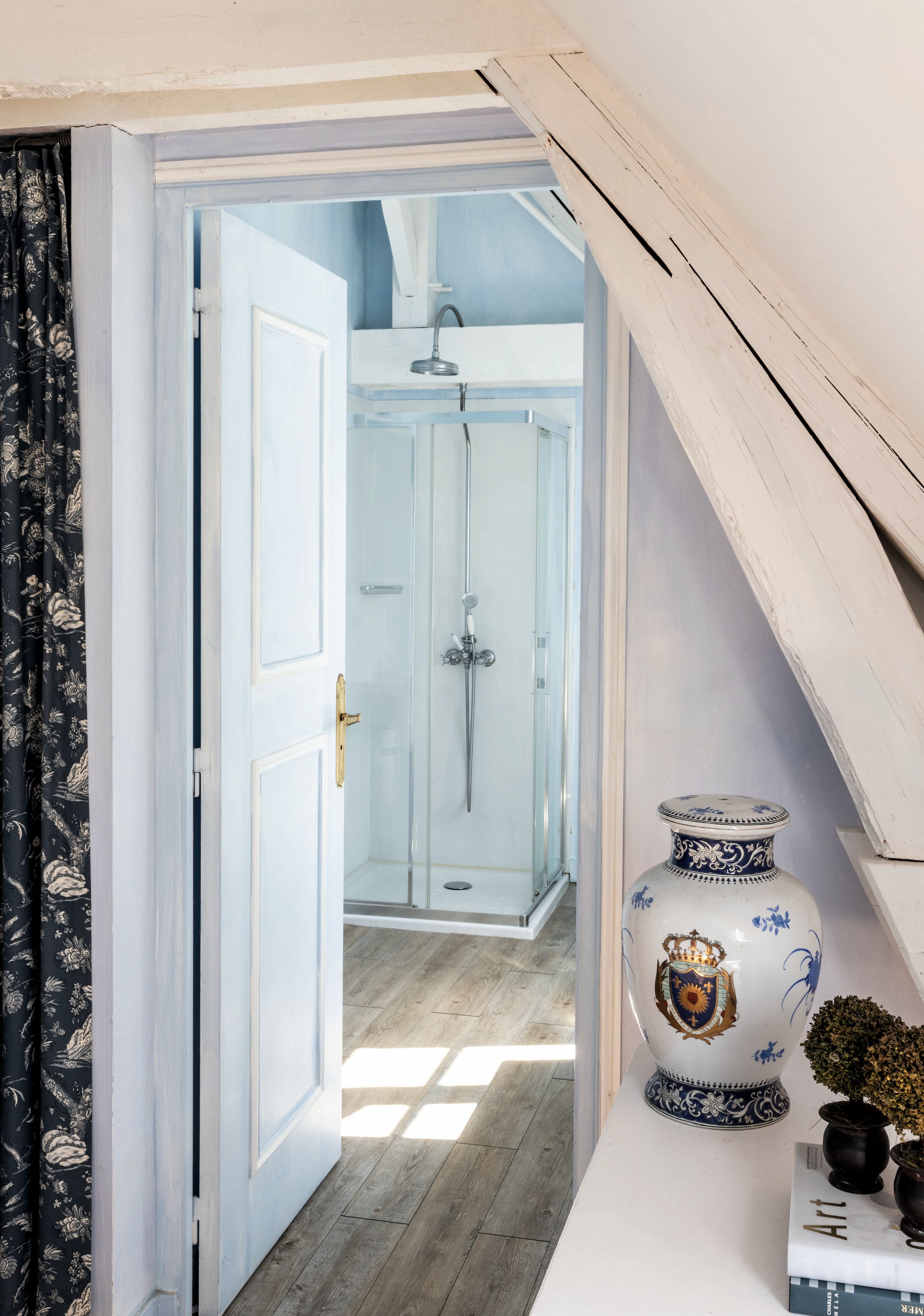 Puerta abierta a un baño con ducha de vidrio, jarrón decorativo y plantas.