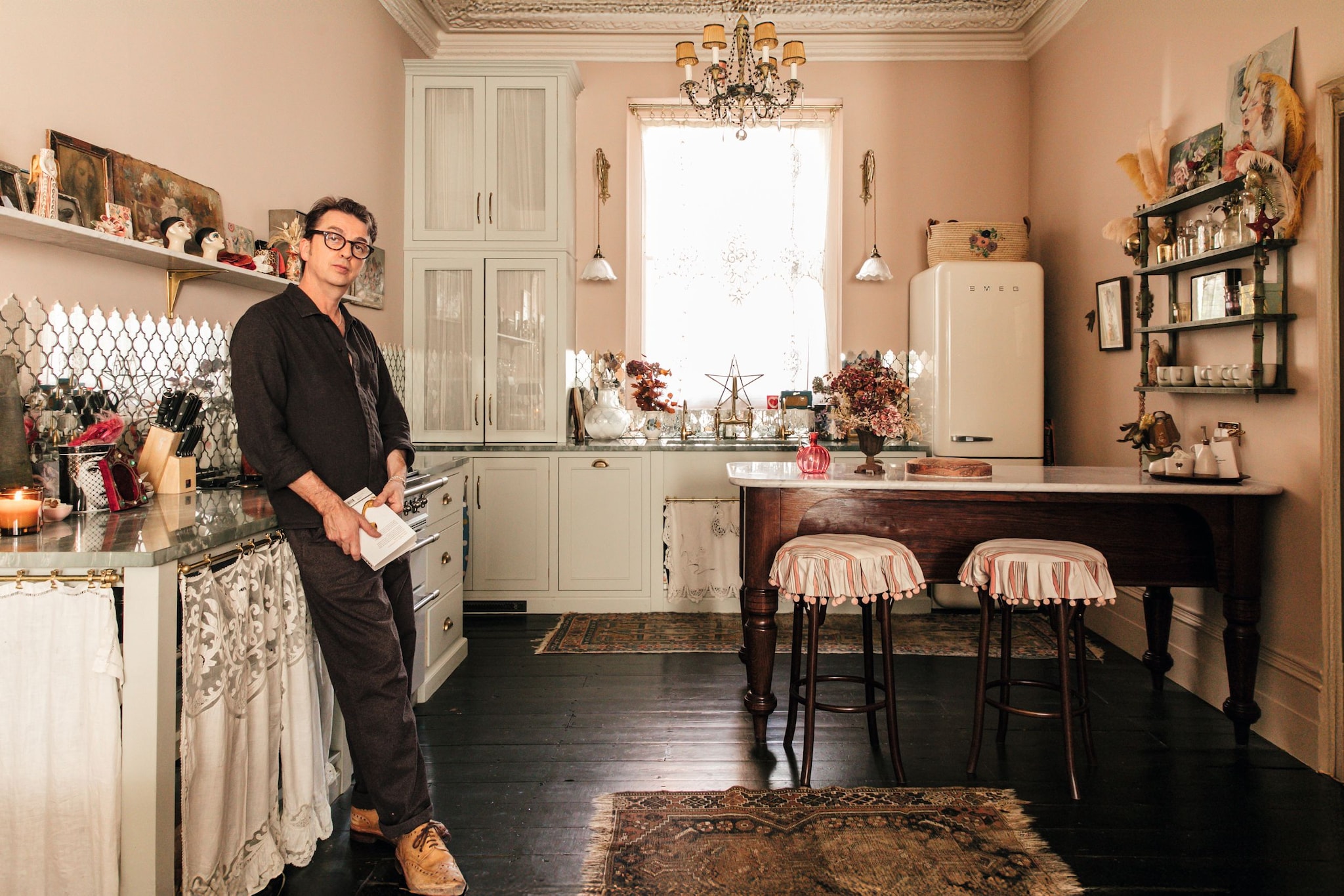 Danny Goffey en una cocina vintage, con camisa negra y pantalones oscuros, sostiene un libro.