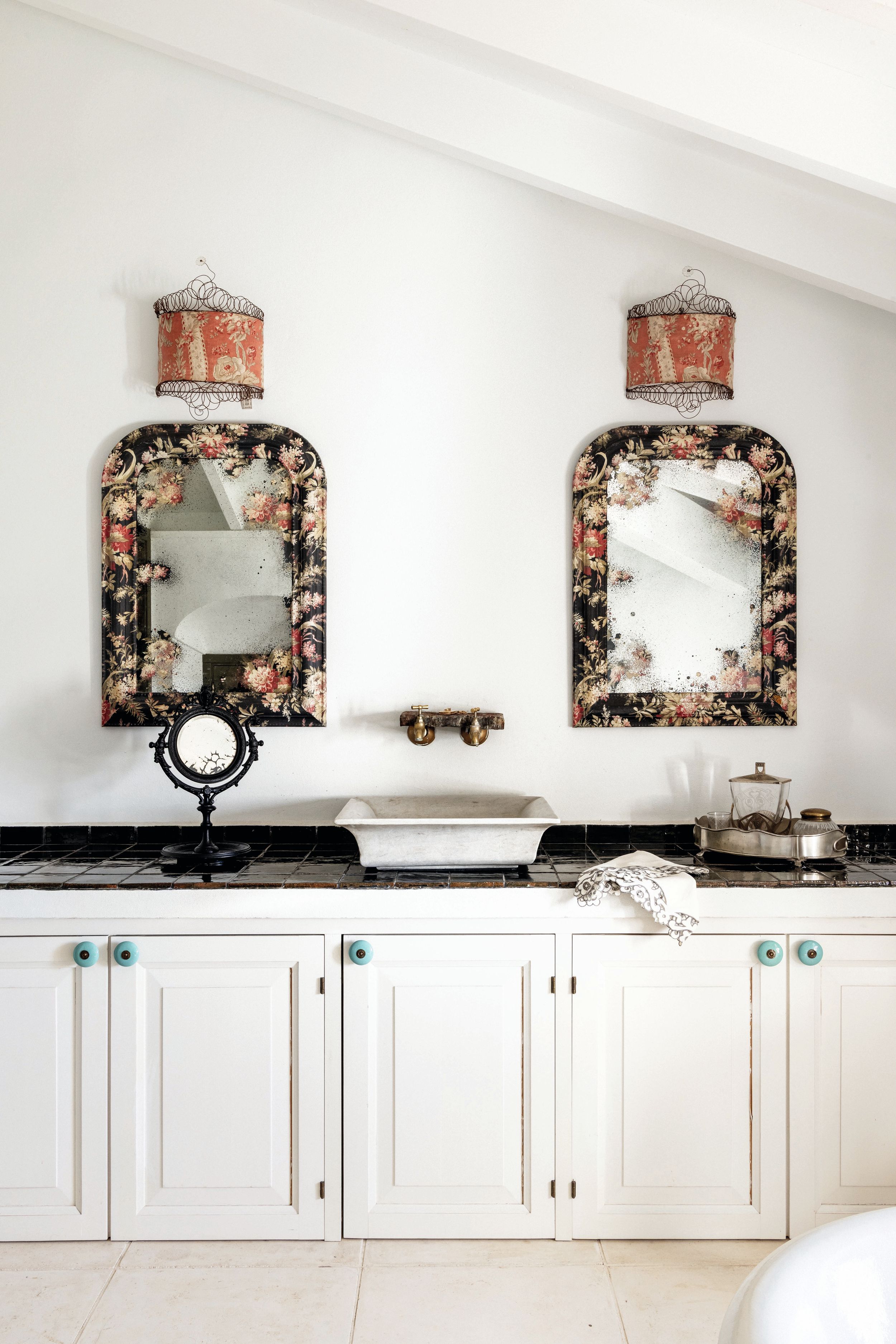Baño elegante con espejos florales, lámparas vintage, y gabinetes blancos.