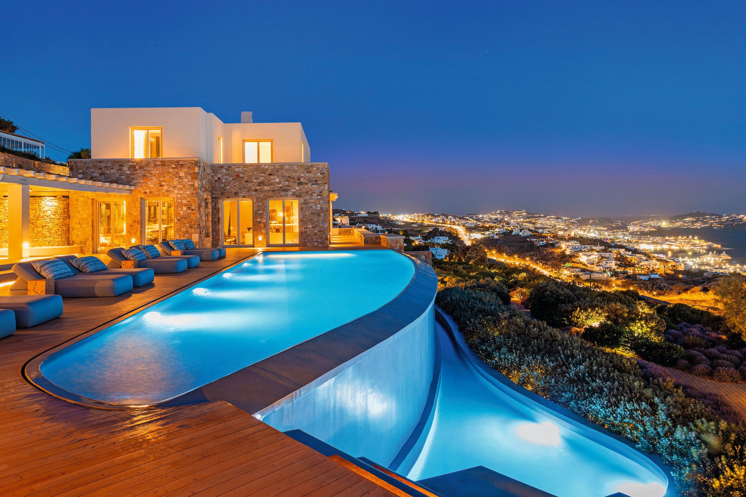 Las dos piscinas infinitas de la villa siguen los contornos naturales de la roca, y desde las amplias terrazas de madera, las vistas panorámicas se extienden a lo largo de la bahía de Mykonos