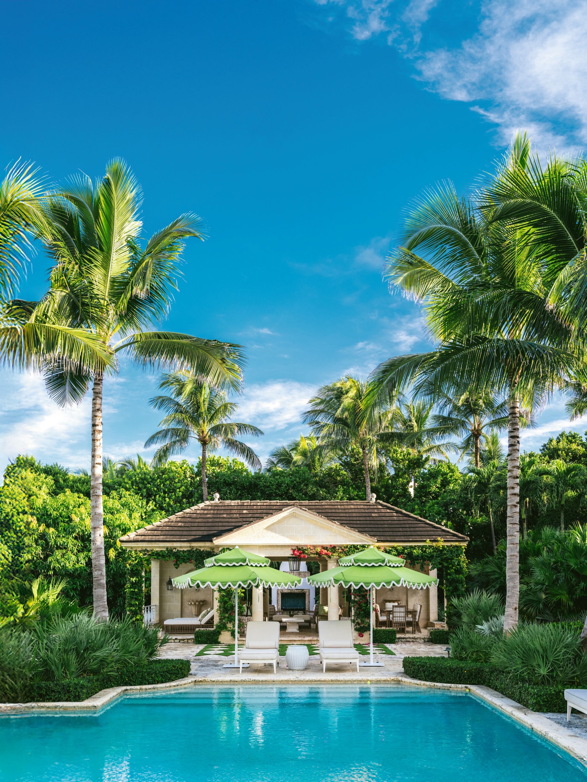 Vista de una casa con piscina, palmeras y tumbones blancas y sombrillas verdes