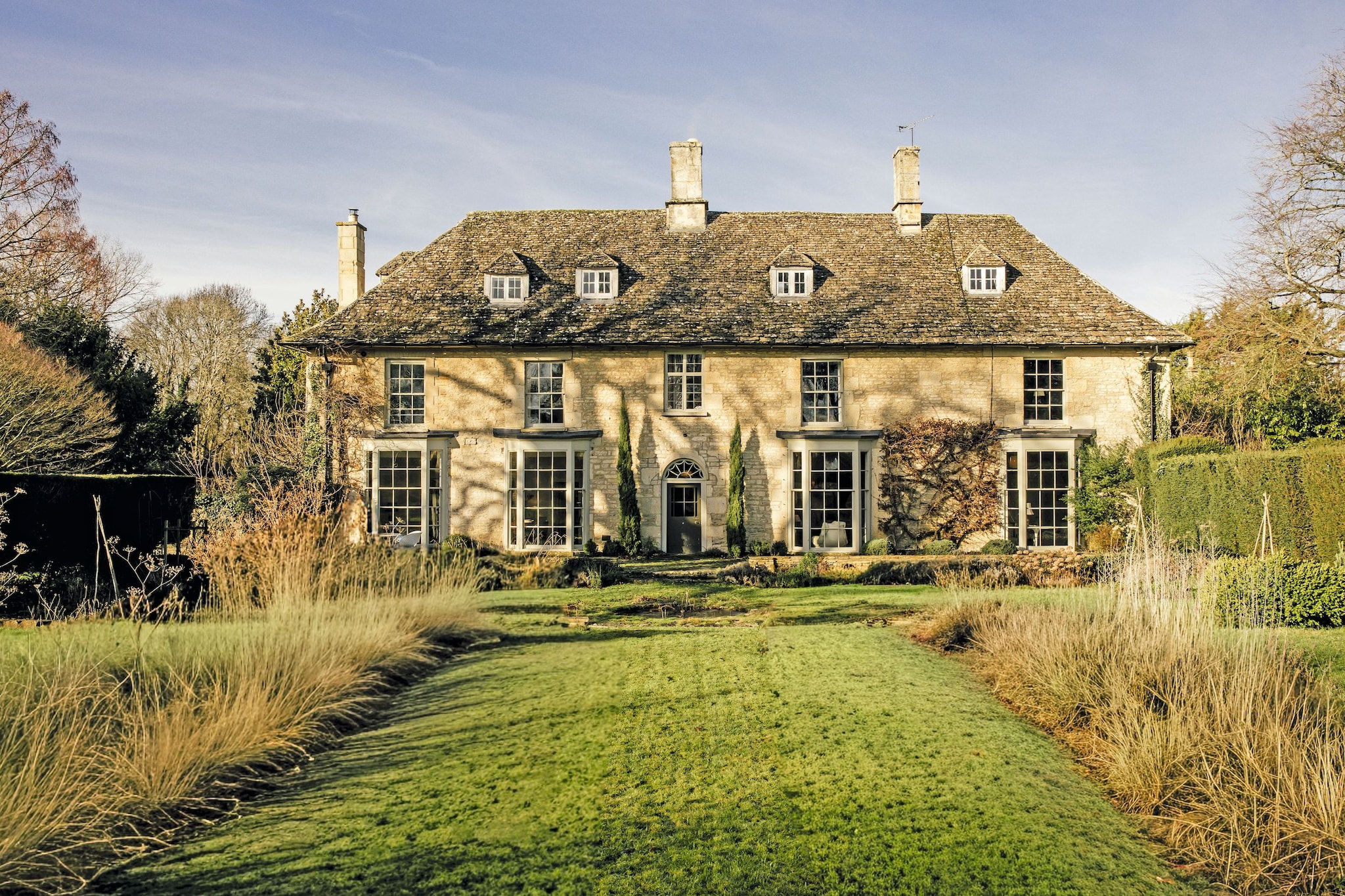 Gran casa de campo con techo de tejas, ventanas y chimeneas, rodeada de jardines.