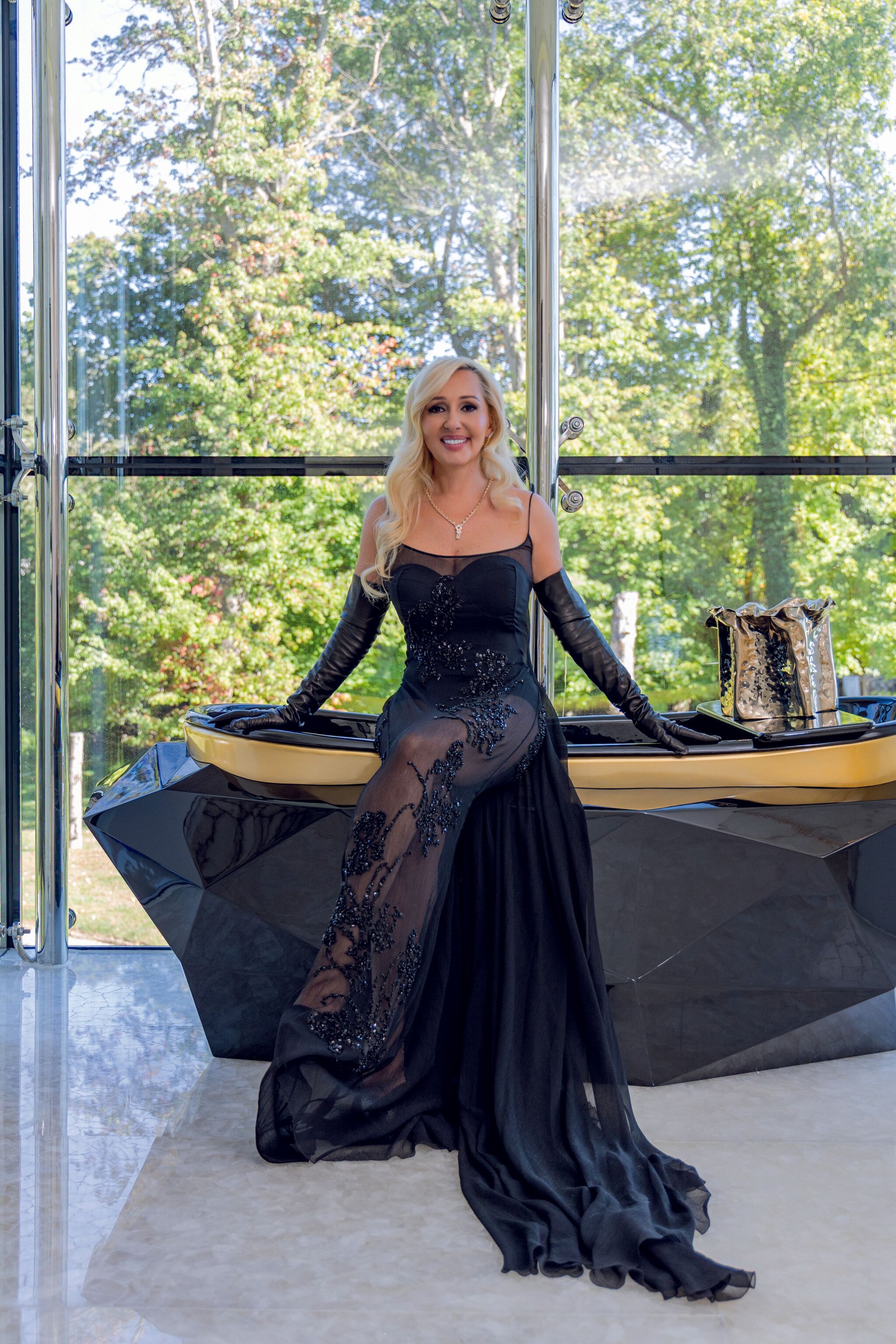 Monique Delacroix en vestido negro posa junto a una mesa de diseño con árboles al fondo.