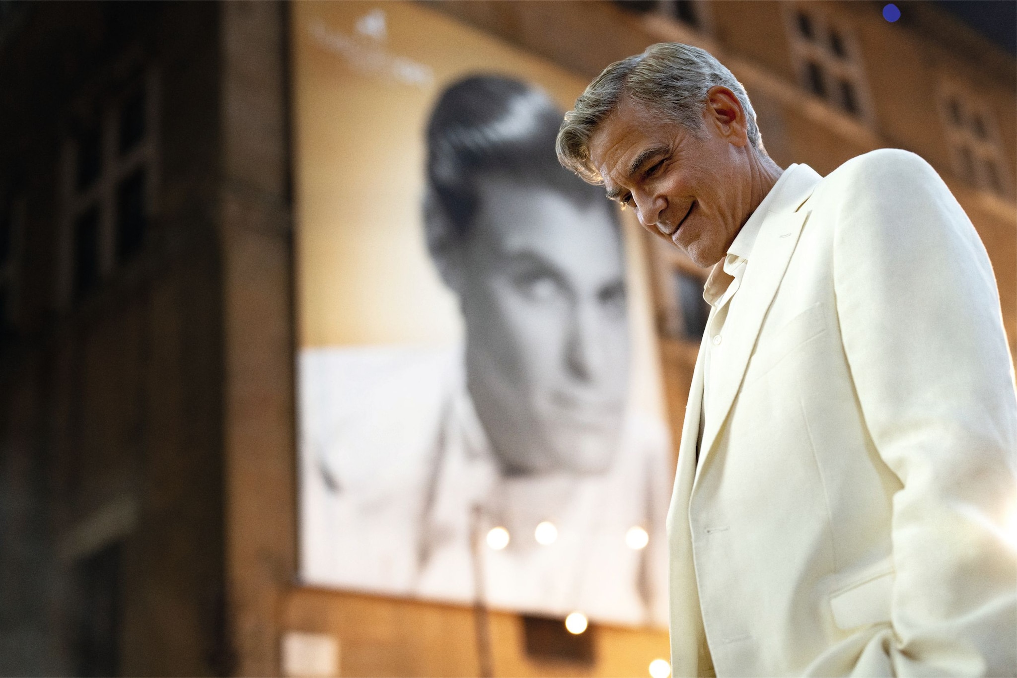George Clooney, en traje blanco, sonríe frente a un póster de un hombre.
