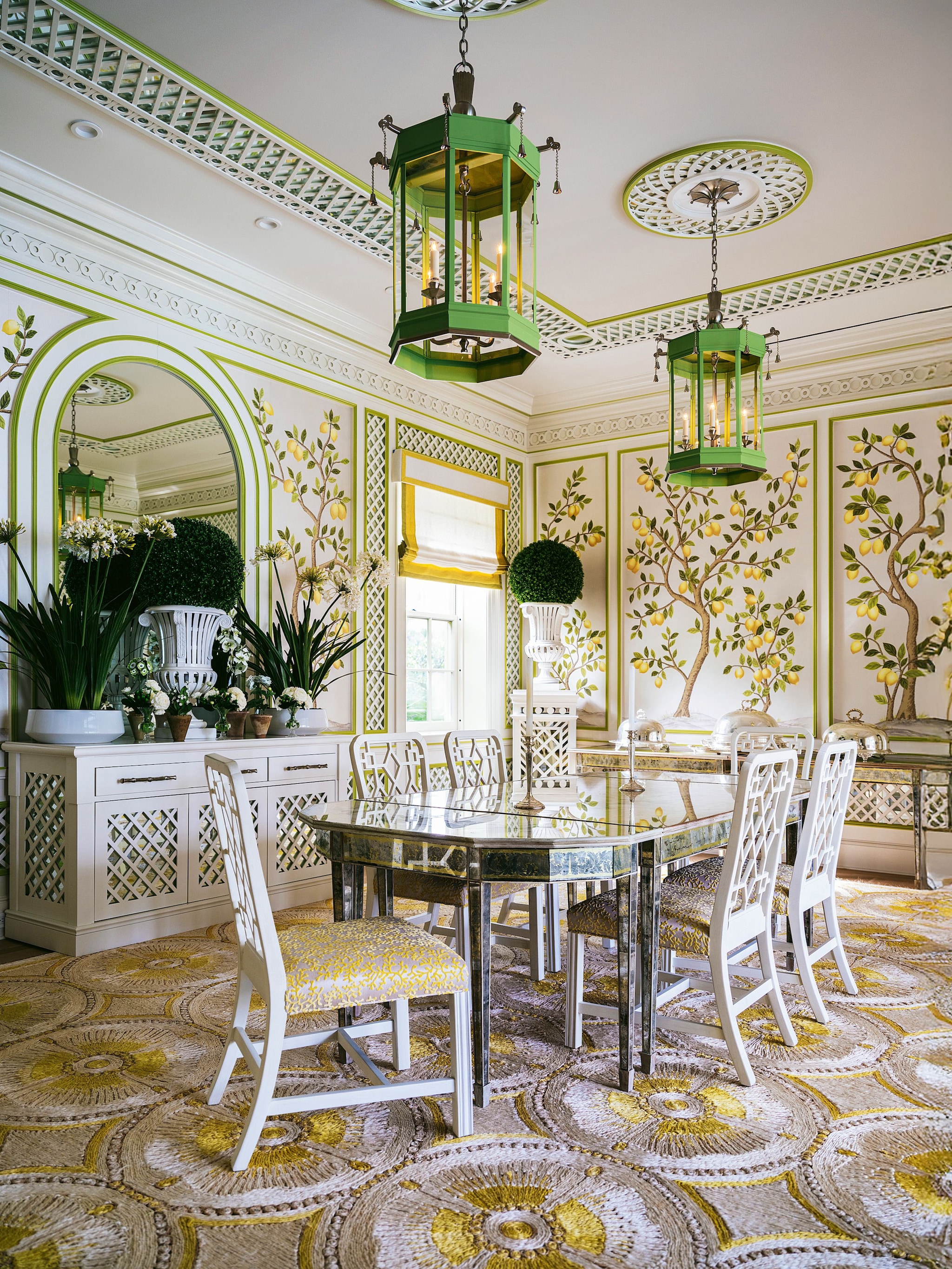 Comedor con paredes con estampados de limoneros, un mesa central de vidrio, sillas blancas y lamparas verdes