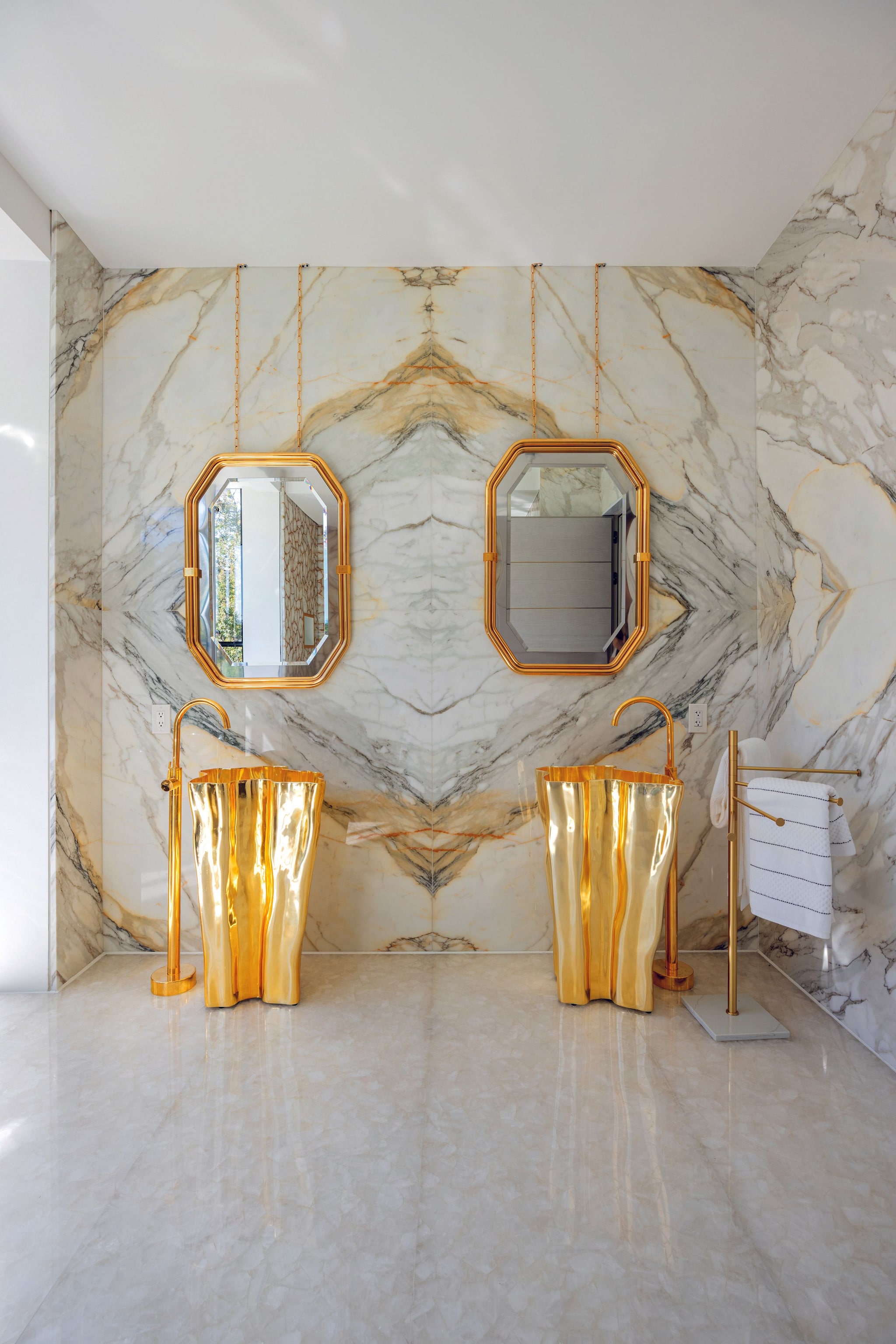 Baño lujoso con dos espejos dorados, lavabos dorados y paredes de mármol.