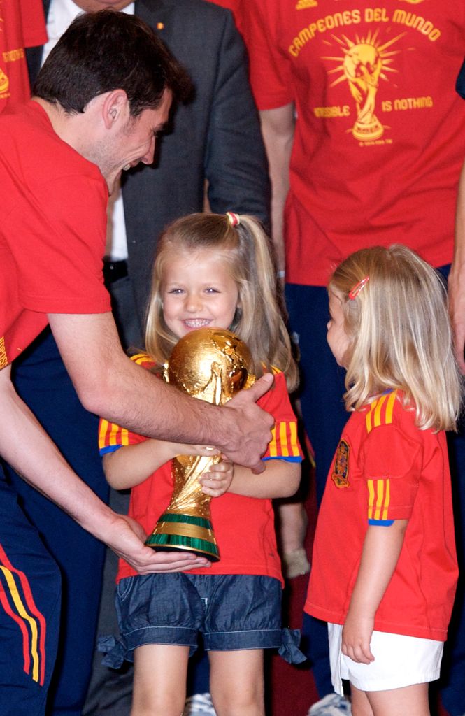 Iker Casillas, el entonces capitán de la selección española, entrega a la princesa Leonor de España la Copa del Mundial, mientras la infanta Leonor contempla el trofeo. Esto se dio en la ceremonia de recepción tras ganar el Mundial Sudáfrica 2010. La imagen fue tomada en el Palacio de la Zarzuela, el 12 de julio de 2010.
