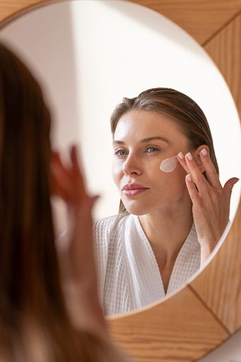 Mujer joven echándose crema en la cara frente al espejo 