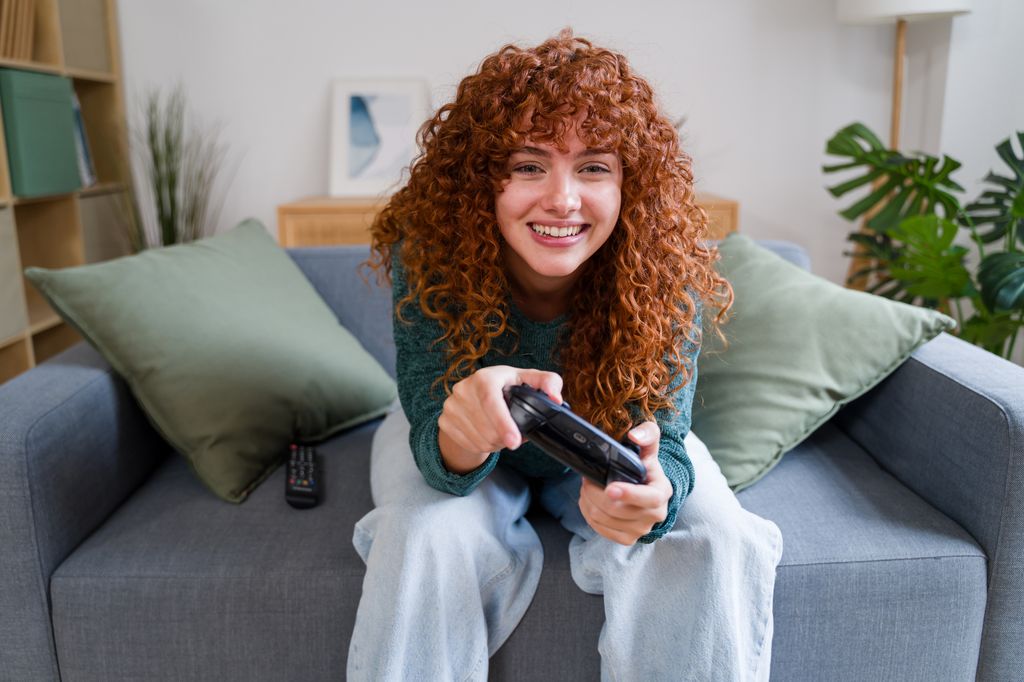 Mujer pelirroja con pelo rizado sentada en el sofá de su casa jugando con una consola