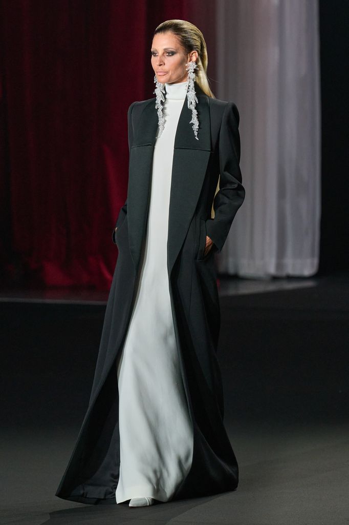 Modelo en pasarela con abrigo negro y vestido blanco, aretes grandes.