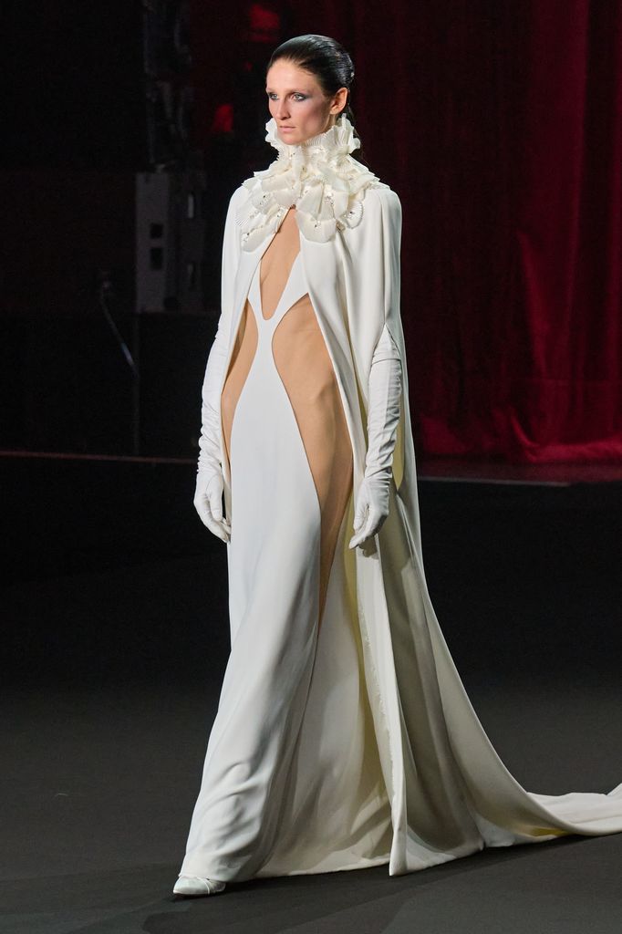 Modelo en pasarela con vestido blanco largo y guantes, cabello negro recogido.