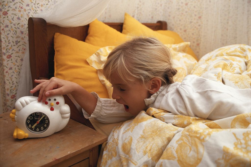 niña en la cama, apagando la alarma del despertador