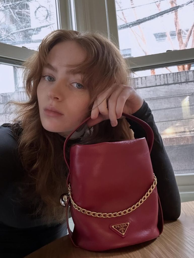 Primer plano de Sadie Sink con flequillo desestructurado, posando junto a un bolso burdeos de Prada con cadena dorada.