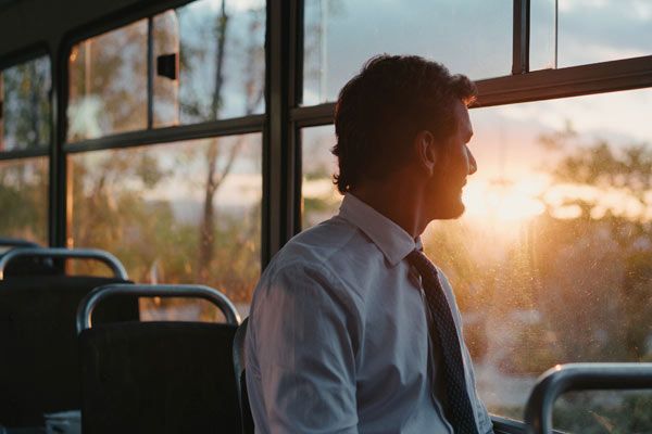 Hombre en traje y corbata sentado en autobús al atardecer, mirando por la ventana.