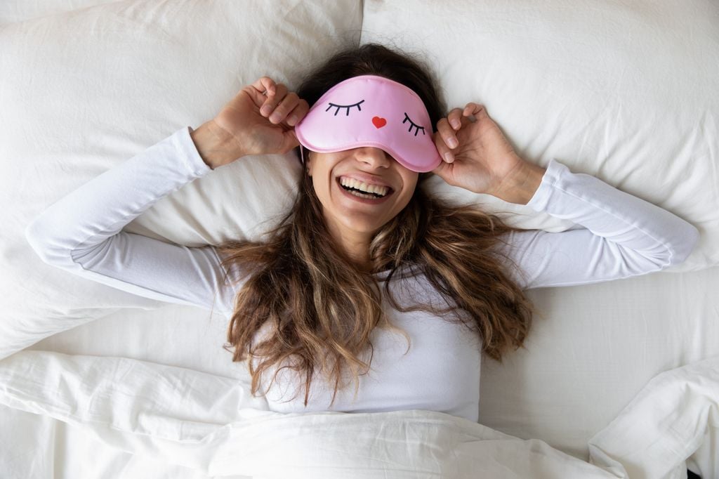 mujer sonriente en la cama, con un antifaz rosa sobre los ojos