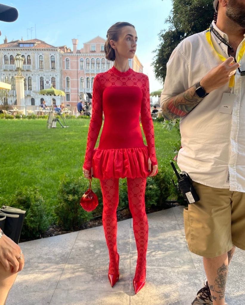 Lily Collins posa con vestido de Gucci rojo con falda globo y medias monogramadas a juego, en un jardín con fondo arquitectónico clásico.