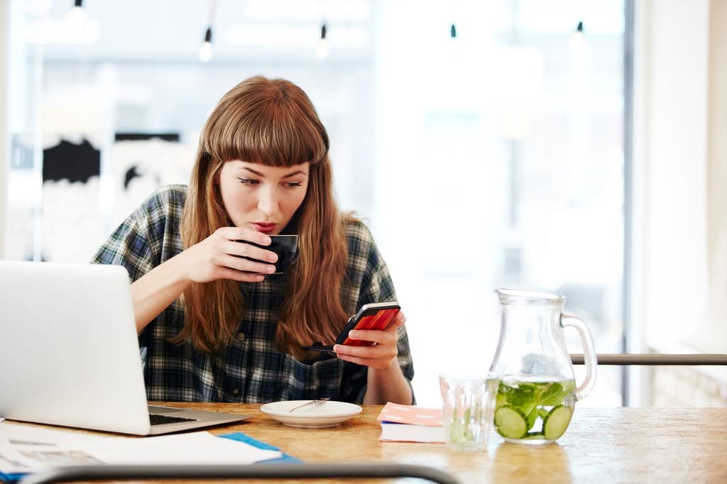 mujer tomando un café mientras mira su móvil y tiene abierto el portátil