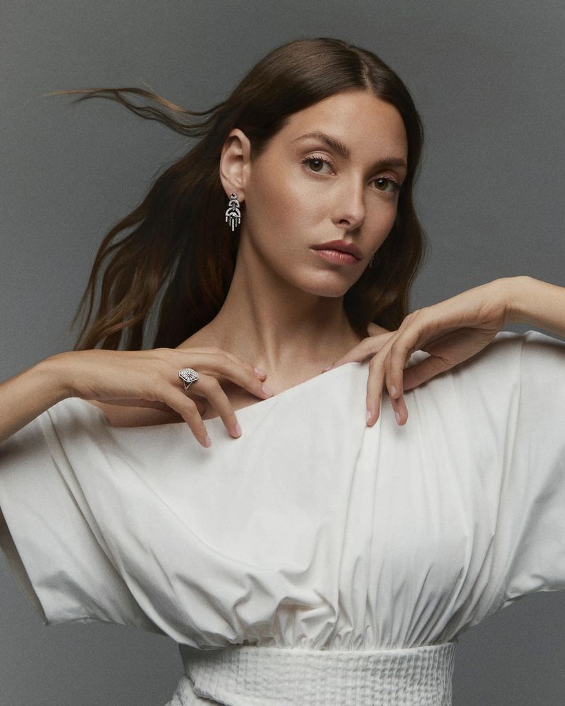 Mujer con cabello largo, vestido blanco y joyas, posando con las manos en el escote.