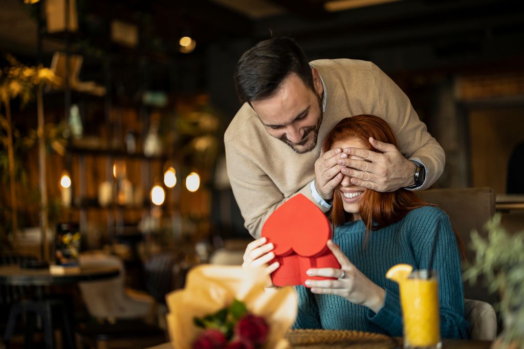 Hombre dando un regalo sorpresa a una mujer con una caja en forma de corazón y tapándole los ojos