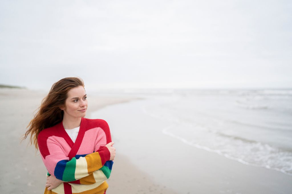 Mujer con un jersey de colores paseando en la playa con gesto sereno