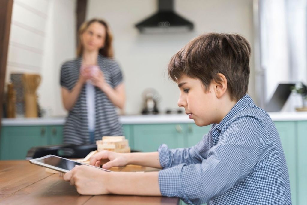 Niño revisando una tablet en la cocina de su casa mientras su madre lo supervisa