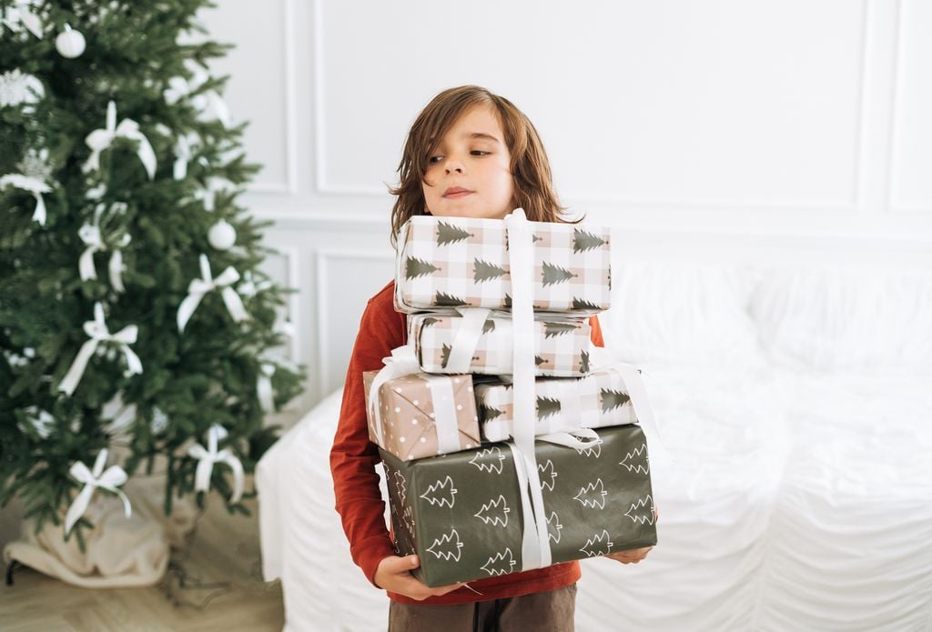 Niño de 6 u 8 años con muchos paquetes de regalos de Navidad en las manos. El fondo es blanco y hay un árbol de Navidad decorado tras él.