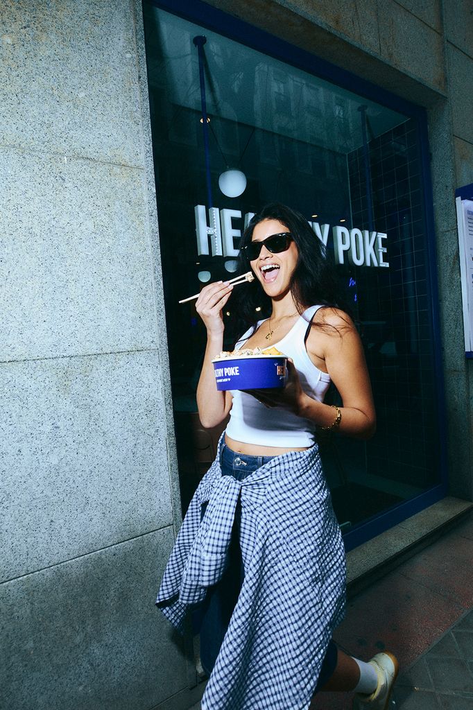 Mujer joven morena comiéndose un Poke bowl de Healthy Poke sonriendo mientras camina por la calle