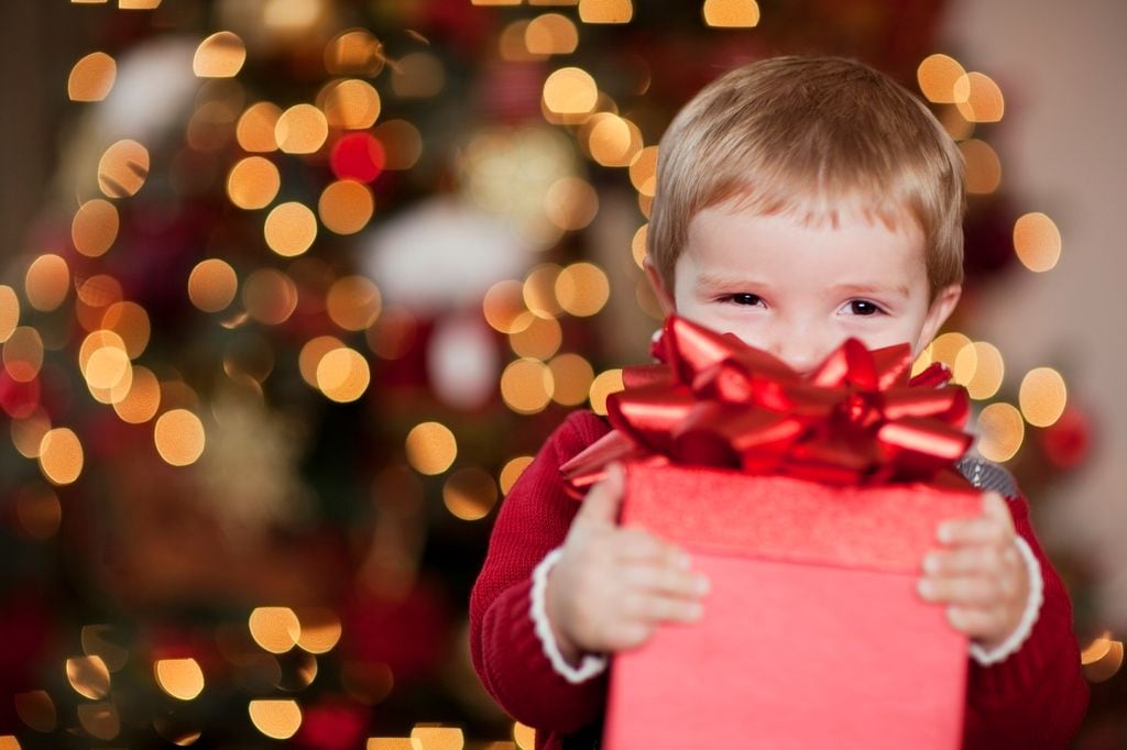 Niño sonriendo detrás de una caja de regalo roja, que tiene con las manos. El fondo está iluminado por Navidad.